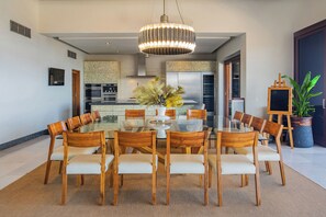 A modern dining area with a glass table surrounded by wooden chairs, a central light fixture, and a kitchen in the background. - 2 Villas, Luxury Amenities & Fully Staffed Service (Cabo San Lucas)