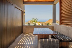 A modern outdoor seating area with a wooden table and benches, a concrete wall, and a view of the ocean and hills. - 2 Villas, Luxury Amenities & Fully Staffed Service (Cabo San Lucas)