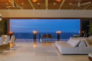 A modern living room with a large glass wall, a sofa, a dining table, and two fire pits. - 2 Villas, Luxury Amenities & Fully Staffed Service (Cabo San Lucas)