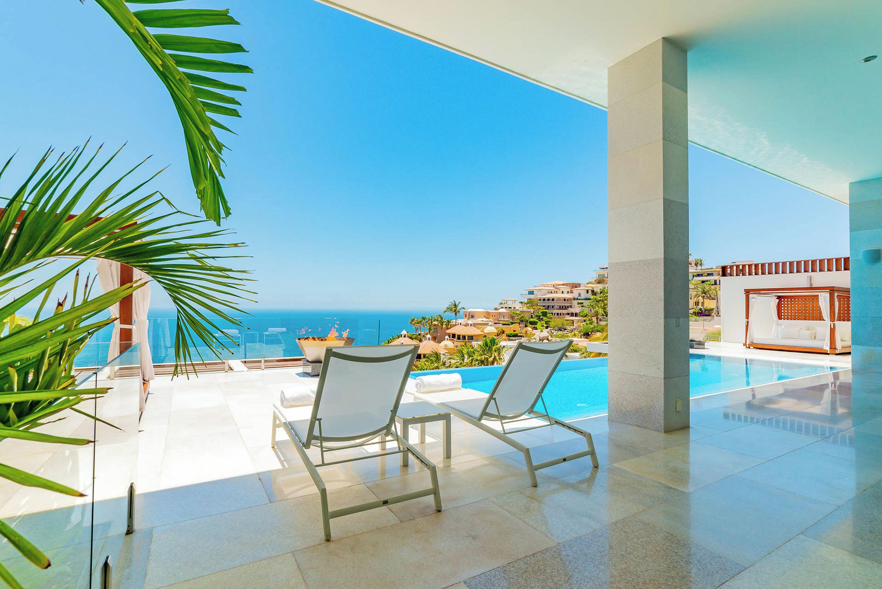 A modern outdoor pool area with two lounge chairs, a fire feature, and a view of the sea and coastal buildings.