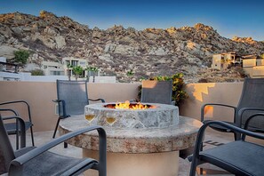 An outdoor seating area with a stone fire pit, two glasses of wine, and a view of a rocky hillside with buildings in the background. - Luxury Pedregal Villa Pairing with Stunning Ocean Views (Cabo San Lucas)
