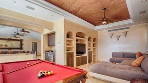 Ein Billardtisch mit Kugeln, eine Couch, ein Fernseher und ein Deckenventilator in einem Raum mit Ziegeldecke.