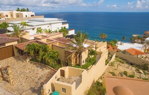 A coastal residential area with terracotta-roofed buildings, palm trees, and a view of the sea. - Luxury Pedregal Villa Pairing with Stunning Ocean Views (Cabo San Lucas)