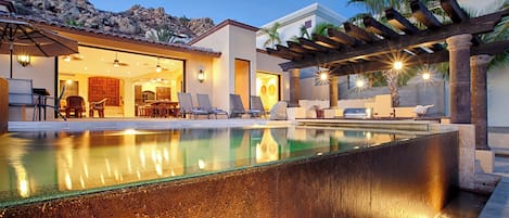 Un área de piscina moderna al aire libre con una pérgola, sillones de descanso y un comedor con vistas a la montaña.