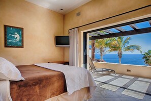 Una camera d'albergo con un letto, una televisione, un quadro e un balcone con vista sul mare e sulle palme.