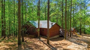 A cabin in a forest with a metal roof, surrounded by trees. - Whispering Pines Retreat - Covered Outdoor Lounge (Broken Bow)