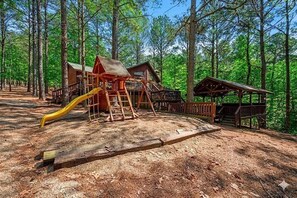 A wooden cabin with a deck and a play structure in a forest. - Whispering Pines Retreat - Covered Outdoor Lounge (Broken Bow)