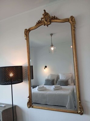Ein Zimmer mit einem großen, verzierten Spiegel, einem Bett mit weißer Bettwäsche, einer Wandlampe und einer Deckenleuchte.