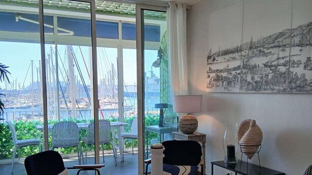 Un salon moderne avec vue sur une marina, comprenant une table ronde, deux fauteuils et une grande fenêtre.