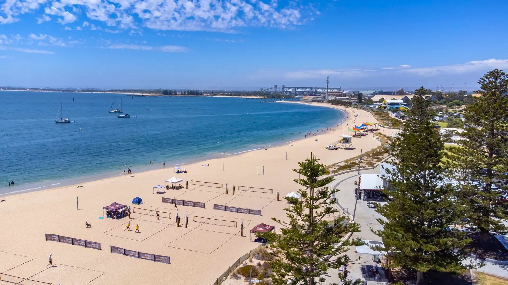 A beach with volleyball courts, a lifeguard tower, and a clear view of the ocean and distant structures.