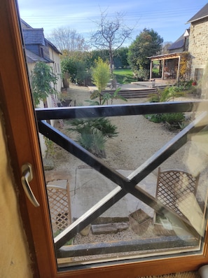Vista a través de una ventana con marco de madera, mostrando un jardín con patio, una mesa pequeña y una estatua.