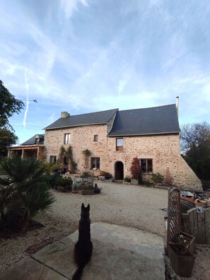A stone building with a sloped roof, a gravel driveway, and a dog in the foreground. - Gîte au Pressoir: Outbuilding on an Old Breton Farm (Corseul)