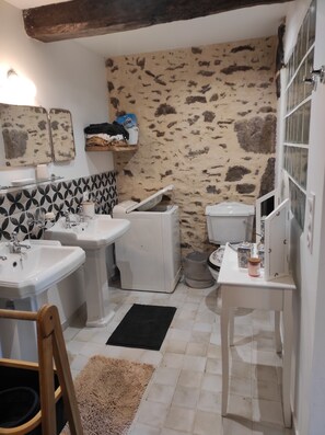 Baño con dos lavamanos, un inodoro y una mesita con un tarro. El salpicadero es de azulejos con diseño y la pared de piedra.
