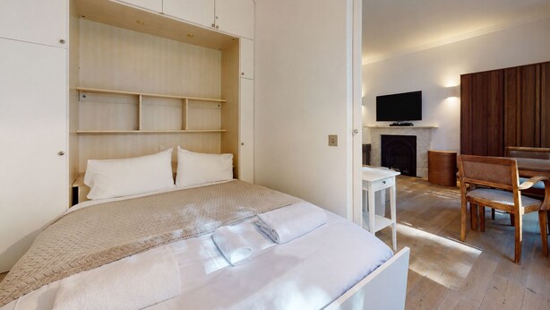 A bedroom with a bed, a TV, a fireplace, and a dining area with chairs. - Bright 1BR Kensington Flat-8 min Walk to Tube (London)