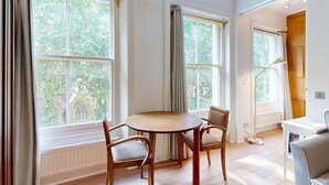 A room with a round table, two chairs, large windows, a radiator, and a wooden cabinet. - Bright 1BR Kensington Flat-8 min Walk to Tube (London)