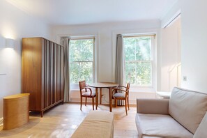 A room with a wooden cabinet, a round table with chairs, a sofa, and two windows with curtains. - Bright 1BR Kensington Flat-8 min Walk to Tube (London)