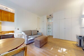 A modern living room with a sofa, ottoman, wooden table, and a trash bin. - Bright 1BR Kensington Flat-8 min Walk to Tube (London)
