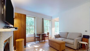 A modern living room with a sofa, ottoman, and a dining table with chairs. There is a fireplace, large windows, and a flat-screen TV mounted on the wall. - Bright 1BR Kensington Flat-8 min Walk to Tube (London)