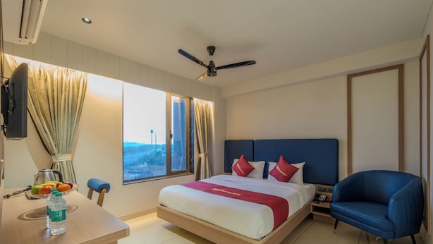 A hotel room with a bed, a desk, a chair, a nightstand, a ceiling fan, and a window with curtains. - The Magnetica (Surat)