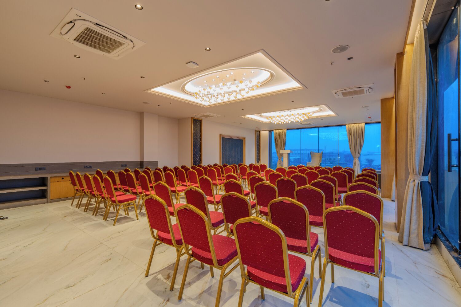 Un salón de conferencias con filas de sillas rojas, una lámpara de araña grande y vistas de la ciudad a través de las ventanas.