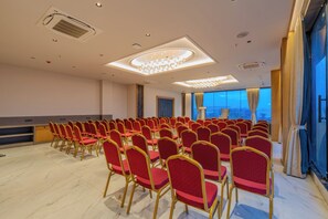 Un salón de conferencias con filas de sillas rojas, una lámpara de araña grande y vistas de la ciudad a través de las ventanas.
