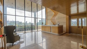 A modern reception area with a wooden reception desk, a grey chair, and large windows offering a view of the cityscape. - The Magnetica (Surat)