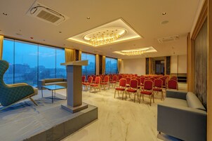 Un salón de conferencias con un atril, filas de sillas rojas, una lámpara de araña grande y vistas de la ciudad a través de amplios ventanales.
