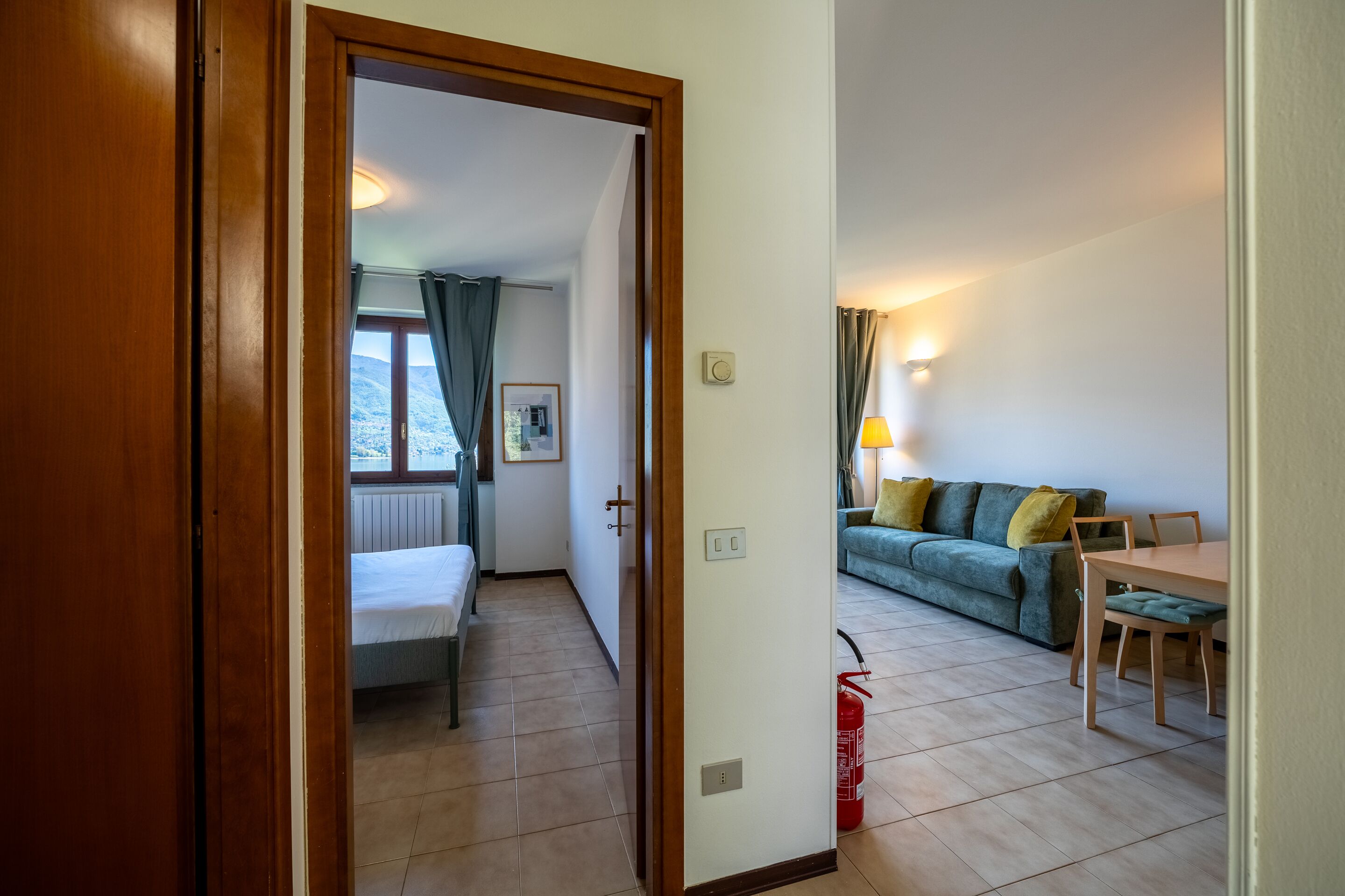Una stanza con un letto, una finestra con tende, una porta, un divano, un tavolo e un estintore.