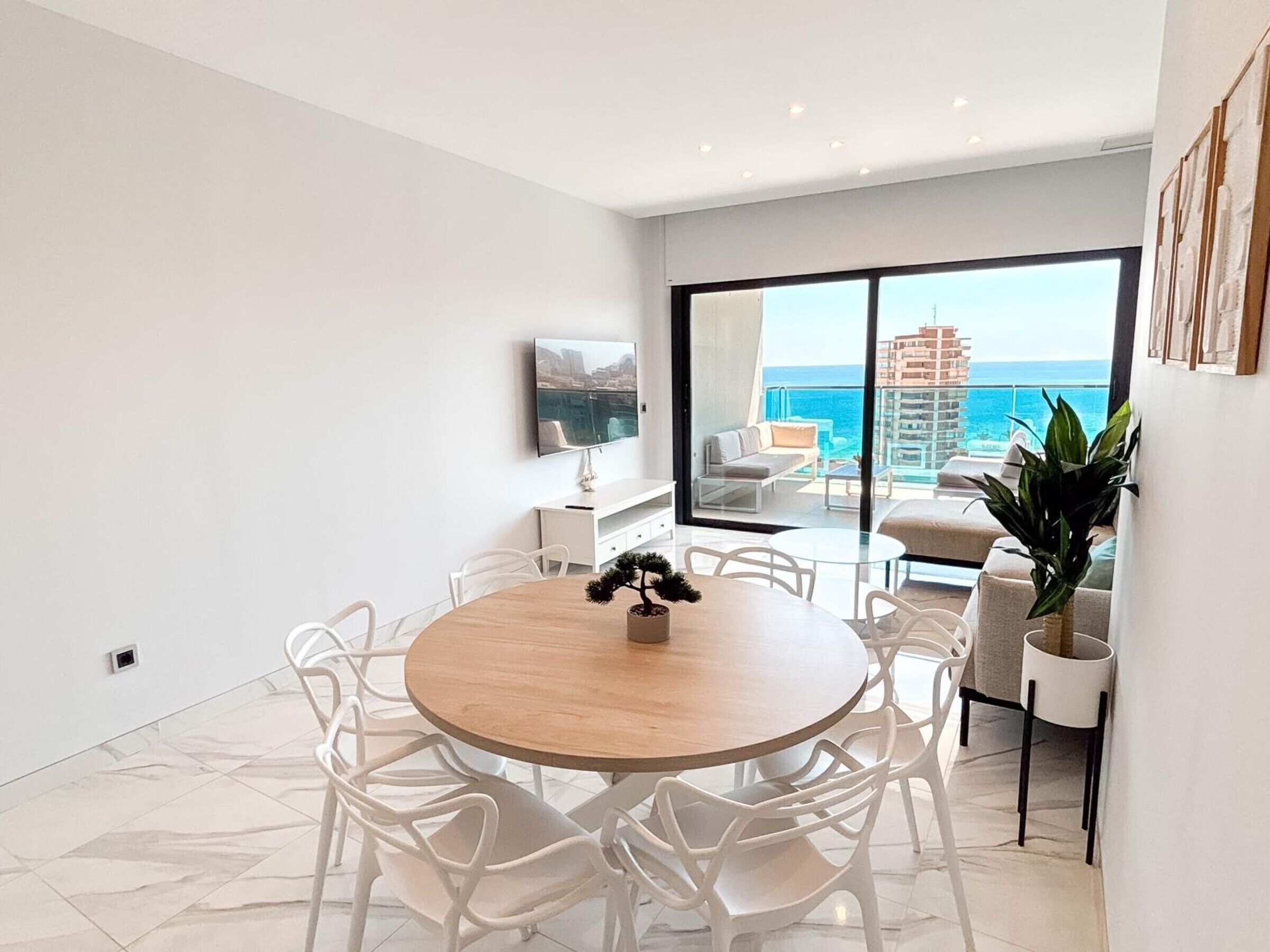 A modern living room with a round wooden table, white chairs, a large window with a view of the sea, and a balcony with outdoor furniture.