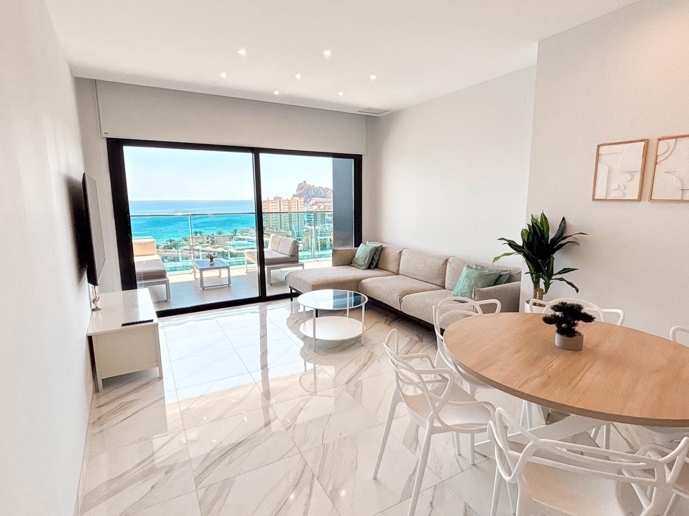 A modern living room with a large sliding glass door leading to a balcony with a view of the ocean and buildings.