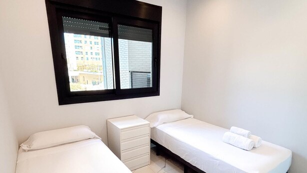 A hotel room with two single beds, a small bedside table, a window with blinds, and a view of buildings outside. - Modern apartment with panoramic sea view (Benidorm)