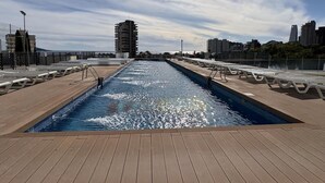 A swimming pool with a wooden deck, surrounded by city buildings and a clear sky. - Modern apartment with panoramic sea view (Benidorm)