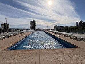 A swimming pool with a wooden deck, surrounded by city buildings and a clear sky. - Modern apartment with panoramic sea view (Benidorm)
