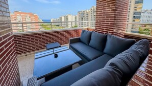 Ein Balkon mit einer Couch und einem kleinen Tisch, mit Blick auf eine Stadtlandschaft aus Gebäuden und einem klaren Himmel.