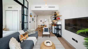 A modern living room with a sofa, a TV, and a kitchen area in the background. - Boho-Chic 1BR w/ Pool | Dubai Hills (Dubai)