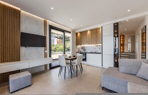 A modern living room with a sofa, a dining table with chairs, a TV mounted on a wall, and a kitchen area with a marble countertop. - Bella Vista Balcony 1BR Apartment (Vlorë)