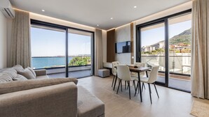 A modern living room with a sofa, dining table, chairs, and a TV. Large windows offer a view of the sea and buildings. - Bella Vista Balcony 1BR Apartment (Vlorë)