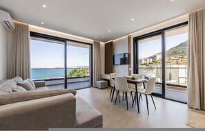 A modern living room with a sofa, dining table, chairs, and a TV. Large windows offer a view of the sea and buildings. - Bella Vista Balcony 1BR Apartment (Vlorë)