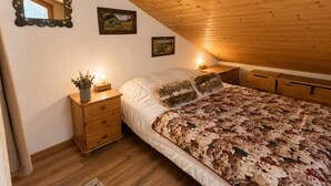 A bedroom with a sloped ceiling, wooden headboard, a bed with a floral-patterned bedspread, two wall-mounted paintings, a bedside table with a lamp, and a mirror with a floral frame. - Spacious duplex, mountain view - 5 guests (Méribel)