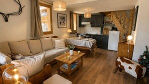 A cozy living room with a sofa, a wooden coffee table, and a lamp. There is a kitchen area with cabinets and a refrigerator. A staircase with wooden railings is visible. - Spacious duplex, mountain view - 5 guests (Méribel)