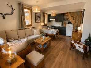 A cozy living room with a sofa, a wooden coffee table, and a lamp. There is a kitchen area with cabinets and a refrigerator. A staircase with wooden railings is visible. - Spacious duplex, mountain view - 5 guests (Méribel)