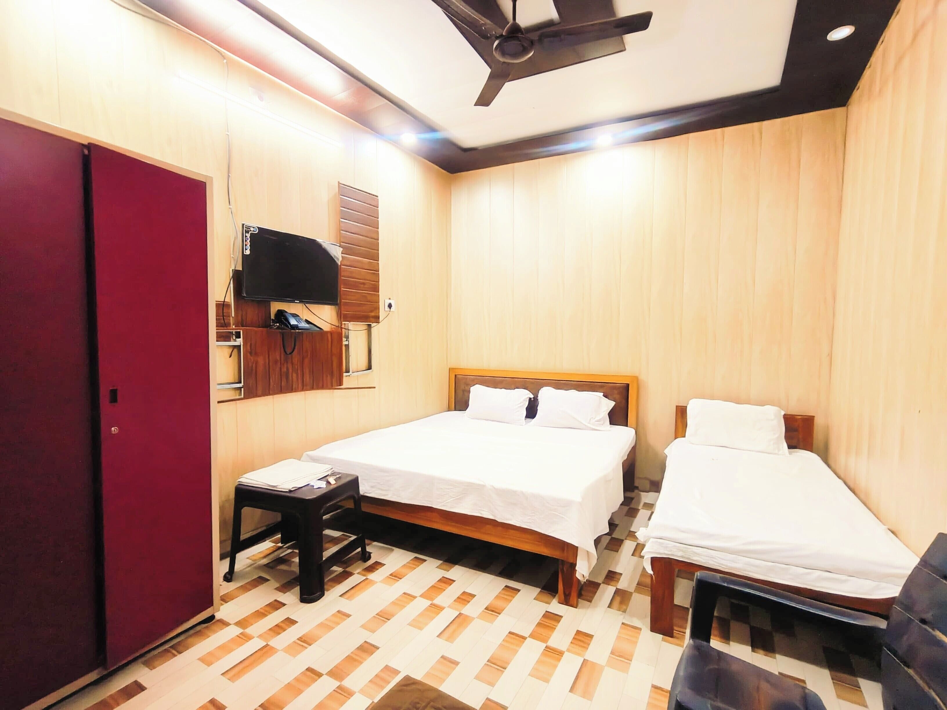 Ein Hotelzimmer mit zwei Betten, einem an der Wand montierten Fernseher, Holzboden und Deckenventilator.