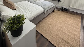 Ein modernes Schlafzimmer mit einem Bett, einem kleinen Nachttisch mit Pflanze, einem Heizkörper und einem Fenster mit Vorhängen.