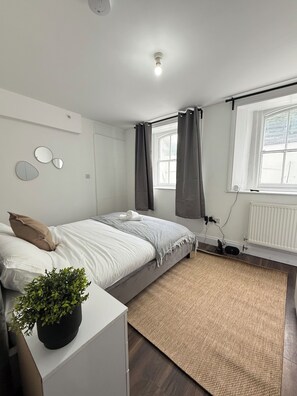 Ein modernes Schlafzimmer mit einem Bett, einem kleinen Nachttisch mit Pflanze, einem Heizkörper und einem Fenster mit Vorhängen.