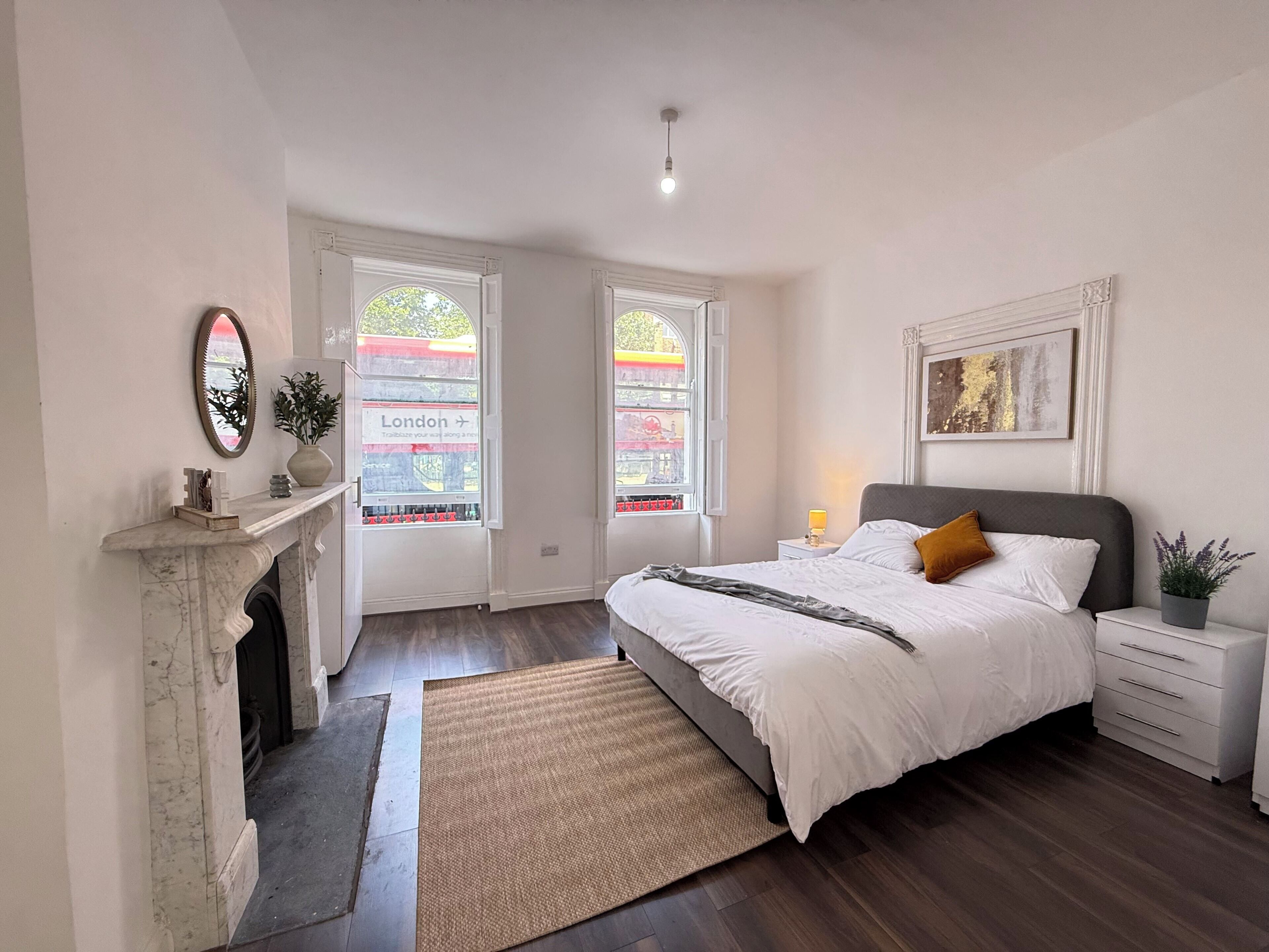 A bedroom with a bed, nightstands, a fireplace, a mirror, and two windows with a view of London.