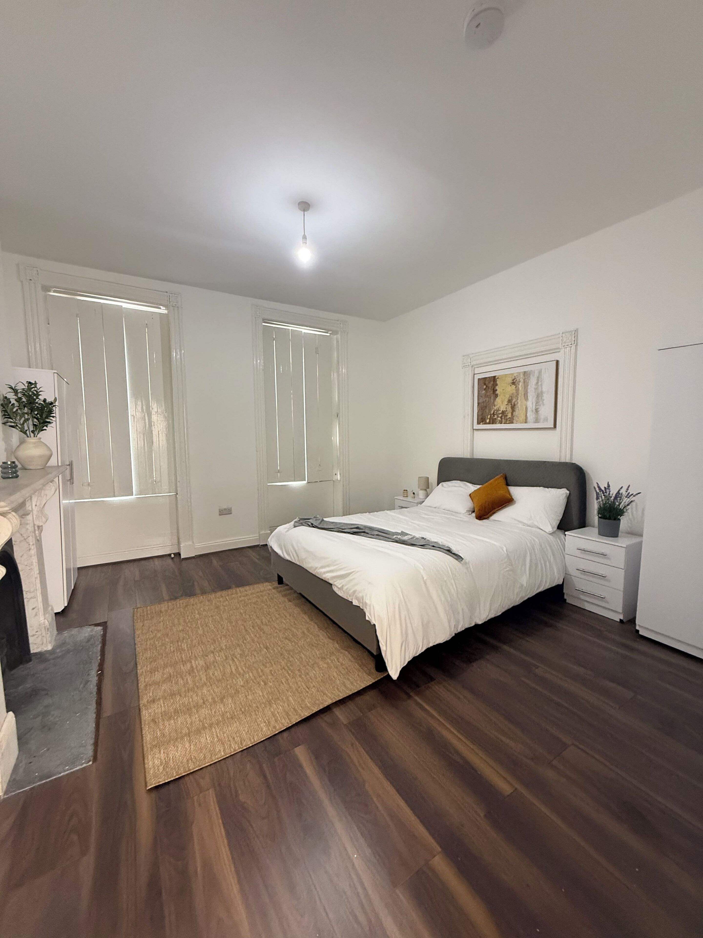 Une chambre à coucher comprenant un lit, des tables de chevet, une commode, une fenêtre avec des stores, une œuvre d’art accrochée au mur et un luminaire au plafond.
