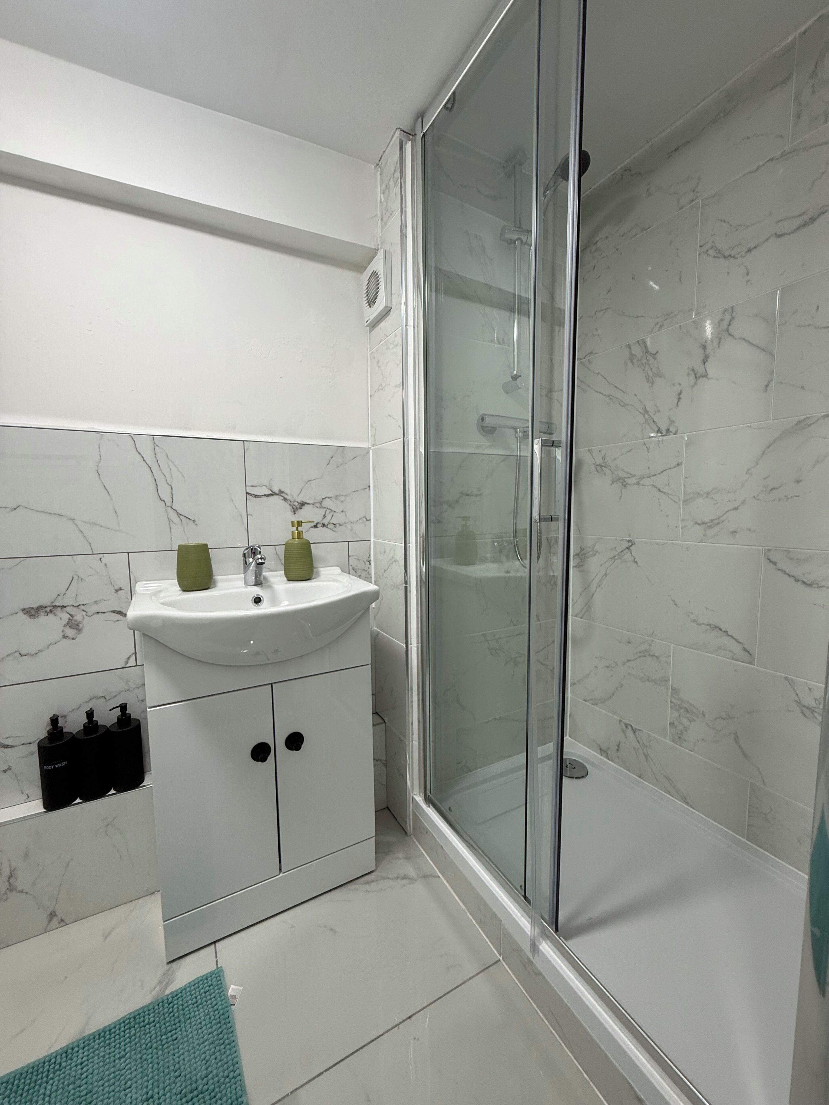 A bathroom with a glass-enclosed shower, a white sink with a green soap dispenser, and marble-patterned tiles.
