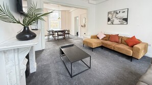 Ein Wohnzimmer mit einer Eckcouch, einem Couchtisch, einem Fernseher, einer Pflanze und einem Essbereich mit Stühlen und einem Tisch.