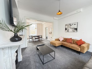 Ein Wohnzimmer mit einer Eckcouch, einem Couchtisch, einem Fernseher, einer Pflanze und einem Essbereich mit Stühlen und einem Tisch.