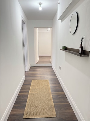 A hallway with a wooden floor, a herringbone-patterned rug, a wall-mounted shelf with a plant and a bottle, and a round mirror. - Amazing Huge 5 Bed / 10 Sleeper in London Zone 2. (London)
